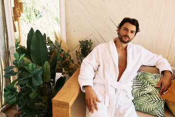 Latin man in white bathrobe rests sitting on couch after baths procedures and wellness at spa resort