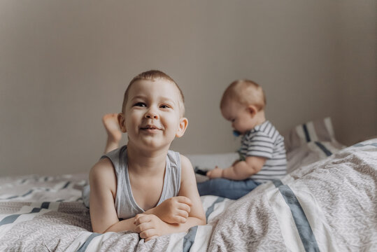 The Two Brothers' Children Are On The Bed. The Older Four-year-old Is Lying And Smiling At The Camera. The Younger One Is Busy Playing With His Phone. He Has A Pacifier In His Mouth.	