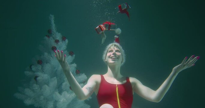 Fun happy attractive young Santa elf fairy woman opens hands with Christmas presents floating up underwater slow motion.