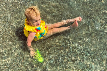Fototapeta premium A little girl toddler is sitting in a vest and a water pistol in the water on the seashore