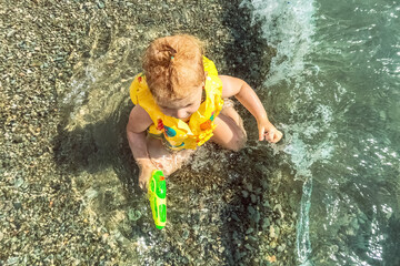 A little girl toddler is sitting in a vest and a water pistol in the water on the seashore