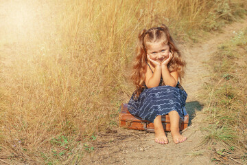 little girl sitting on an old suitcase on a field road