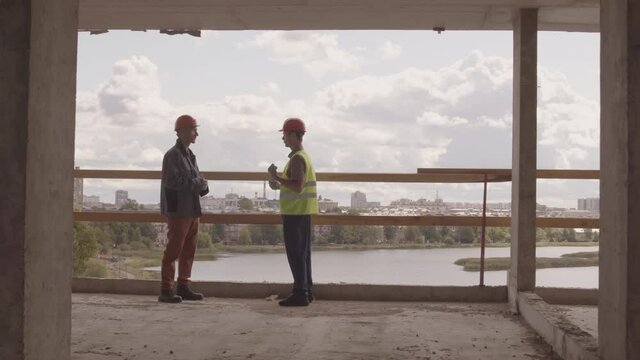 Wide tracking left of two Caucasian workmen wearing helmets standing on edge inside building under construction, talking on summer day