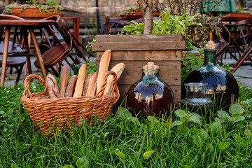 Wicker basket full Of Baguettes on green grass in park, near large bottles with wine.