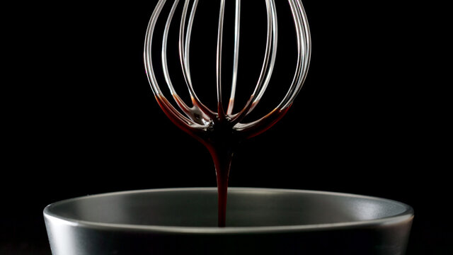 Liquid chocolate dripping from steel whisk on black background