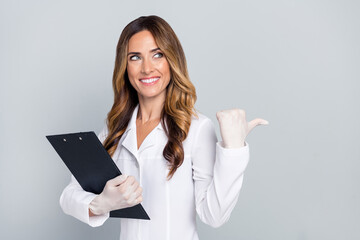 Photo of young woman point finger thumb empty space clipboard wear uniform white coat isolated on grey color background