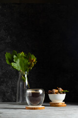 Glass cup with double bottom with coffee on a wooden stand with a plate with an acorns and a glass vase with oak leaves on a dark background