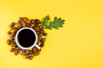 White mug with an alternative drink without caffeine surrounded by ripe acorns with a green oak sheet on a yellow space copy background for text