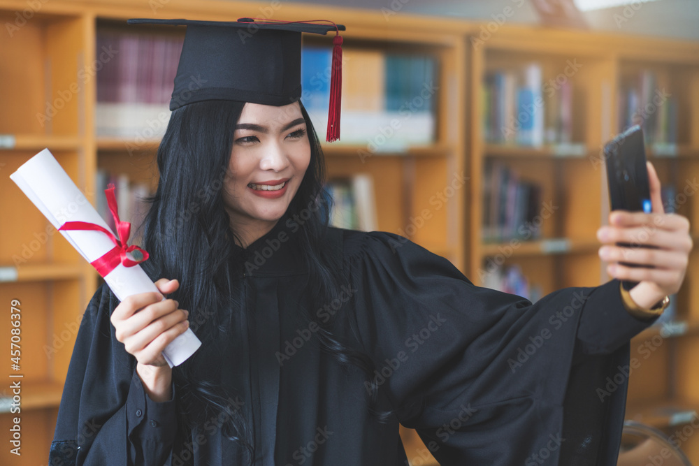 Wall mural University graduate in graduation gown and mortarboard celebrates in a virtual graduation ceremony. Happy female student on her graduation day at home. Concept of online education