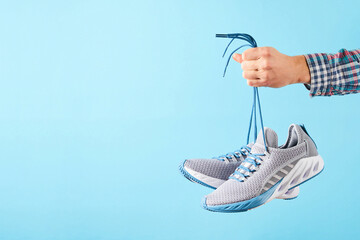 Hand holds hanging gray running sneakers by the laces on blue pastel background. Hand with a new sport shoe. Stability and cushion running shoes.