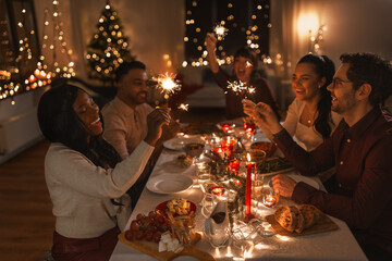 holidays and celebration concept - multiethnic group of happy friends with sparklers having christmas dinner at home