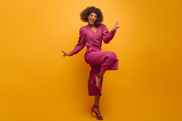  pretty artistic lady in jump shows victory sign with two hands on yellow background. smiling dark-skinned girl in festive lilac dress and pink high-heeled shoes.