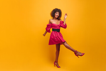  feminine striking African steps forward with beautifully straight leg against backdrop of studio wall. woman with dark brown afro hair is dressed in fashionable crimson dress and open shoes.