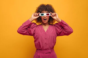 closeup shocking african woman holding 3D glasses on her face with two hands on isolated background. young curly-haired brown-haired woman with open mouth is dressed in festive lilac outfit.
