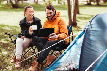 camping, tourism and travel concept - happy couple with tablet pc computer drinking tea at tent camp