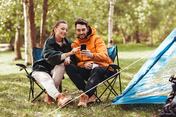 camping, tourism and travel concept - happy couple with smartphone drinking tea at campsite
