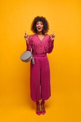 in full growth, sophisticated dark-skinned lady shows with her index fingers up with open mouth. feminine brown-haired woman with lush hair is dressed in loose suit and open shoes in studio.