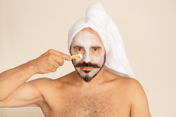 Caucasian man looks at you with face mask and towel on head. White mask is applied with brush