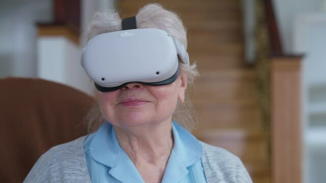 Close-up portrait of happy senior woman in VR googles singing and smiling at home in living room. Happy relaxed Caucasian retiree enjoying karaoke in augmented reality. Modern technologies convenience