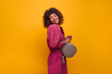  mysterious pretty lady stands half sideways with her eyes closed on yellow background. dark-skinned beauty smiles broadly with teeth dressed in pink outfit and with bag in her hands.