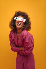  pretty playful girl in 3D glasses smiles broadly with her arms crossed in front of her. curly-haired brunette with afro hair dressed in bright lilac suit against yellow wall.