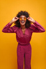 cheerful attractive african woman bent forward holding sunglasses on her face with two hands. young woman with closed eyes smiles broadly dressed in lilac spacious suit.