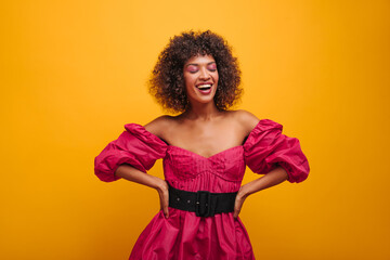 charming cute african smiles broadly with closed eyes indoors studio. young woman holds her waist with both hands and is dressed in crimson dress with black wide belt.