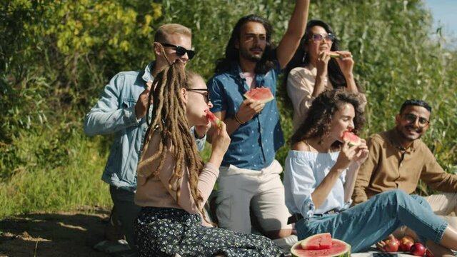 Joyful friends multi-ethnic group are eating watermelon talking and dancing having fun outdoors in park on sunny summer day. Youth and leisure time concept.