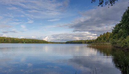 Fototapeta premium Sunny day on a calm river in summer. The blue sky is reflected in the water. Landscape with a lake and a strip of forest on the horizon.