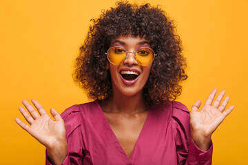 close-up of beautiful African woman in enthusiastic mood on yellow background. curly-haired brunette in translucent glasses with open mouth waving her arms to sides.