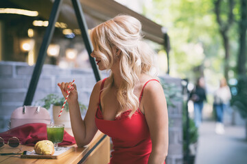 Gorgeous woman in a sexy red dress drinks coffee with a croissant in a cafe on the street.