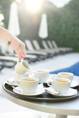 Hand pouring milk into cups with coffee on the table by the pool in the early gentle morning.