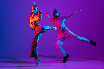 Two dancers, young man and woman dancing hip-hop in casual sports youth clothes on gradient purple pink background at dance hall in neon light. - obrazy, fototapety, plakaty