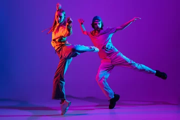 Schilderijen op glas Two dancers, young man and woman dancing hip-hop in casual sports youth clothes on gradient purple pink background at dance hall in neon light. © master1305
