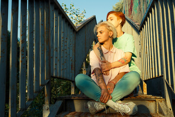 Adorable young lesbian couple in stylish clothes sits together on metal stairs under clear blue sky at sunset in summer evening