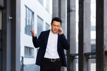 angry and serious Successful Asian businessman explains information to employees using phone, speaks near office outside