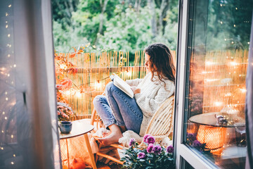 Woman relaxing on cozy balcony, reading a book. - obrazy, fototapety, plakaty