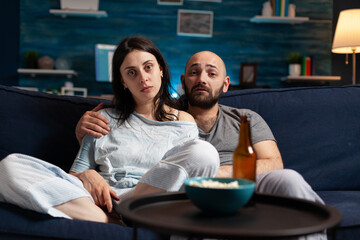 Confused astonished young couple watching documentary show having shocked facial expression, eating popcorn sitting on couch. Concentrated adults looking at tv late night enjoying free time