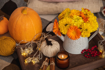 Cozy autumn atmosphere of the house. On a plaid blanket is a wooden tray with decor: a bouquet of yellow flowers, a candle, cinnamon, knitted pumpkin, garland.