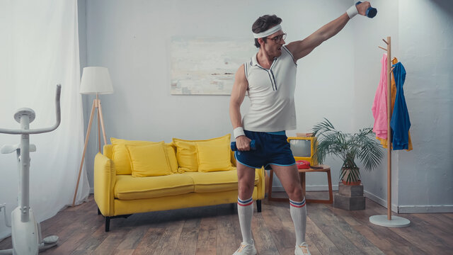 funny sportsman in eyeglasses exercising with dumbbells in living room, retro sport concept