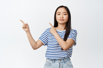 Disappointed asian woman frowning upset, pointing fingers left at advertisement, bad promotion, express dislike and regret, standing over white background