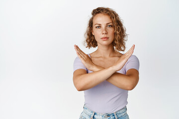 Serious young woman shows cross stop gesture, looking unamused, saying no, forbid something, standing over white background