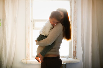Pregnant caucasian mother with long hair with a toddler baby in her arms on the background of window in a lifestyle interior
