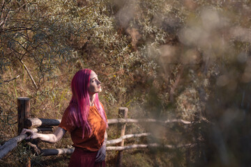 a woman with pink hair stands near a wooden fence in the forest and enjoys the sun