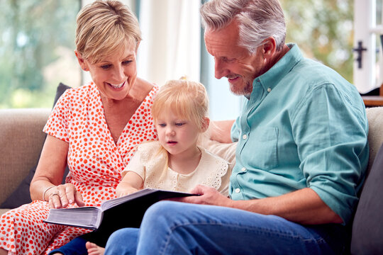Grandparents Sitting On Sofa With Granddaughter At Home Reading Book Together