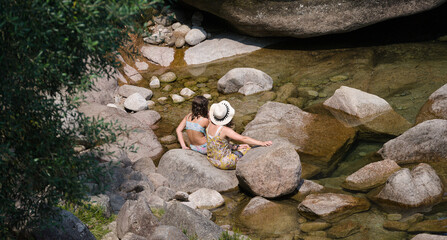 Duas pessoas sentadas num rio de montanha - uma mulher e uma menina - família - chapéu, vestido e...