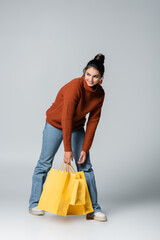 full length of cheerful young woman in sweater and jeans holding yellow shopping bags on grey