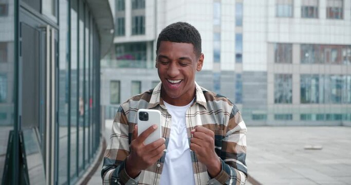 Excited young african american man rejoices at message on his smartphone. Happy male looking at cell phone celebrate good news victory success. Portrait of the lucky gambler showing yes winner gesture