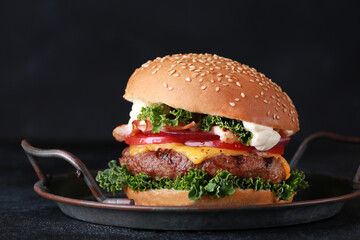 Fast food. Burger with beef, cheese, bacon and vegetables: tomato, onion, cabbage and sauce on a black plate on a black table. Background image, copy space