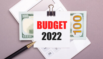 On a gray background, a white calculator, a pen, banknotes and a sheet of paper under a black paper clip with the text BUDGET 2022. Business concept